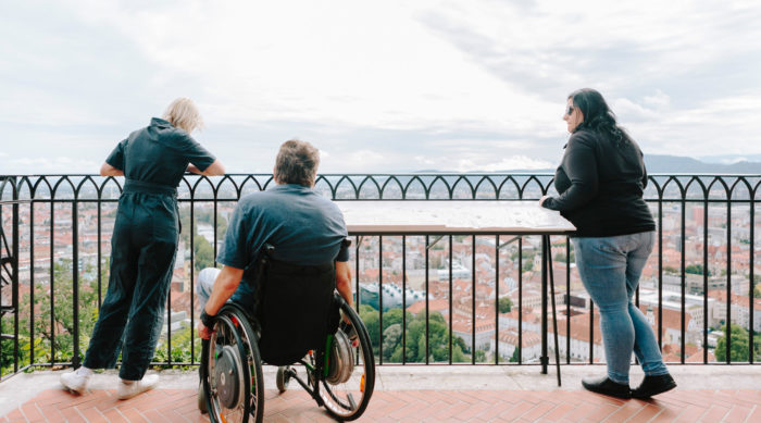 Zwei Frauen und ein Mann im Rollstuhl schauen von der Aussichtsplattform in der Kanonenhalle auf die Stadt Graz herunter. Sie sind von hinten zu sehen.