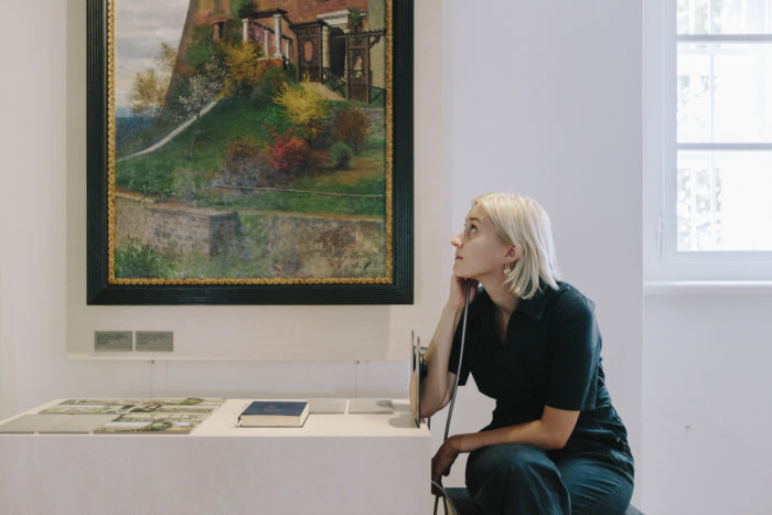 Eine junge Frau sitzt auf einem Hocker an einem Tisch mit Exponaten und betrachtet ein Gemälde im Geschhits-Parcours. Sie hält einen Lautsprecher an ihr rechtes Ohr.