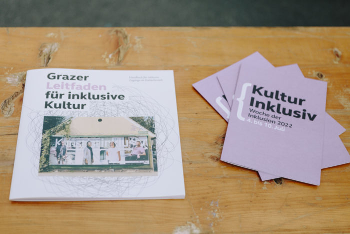 Auf einer Tischplatte aus Holz liegt eine Broschüre mit dem Titel Grazer Leitfaden für inklusive Kultur und drei Folder Kultur Inklusiv.