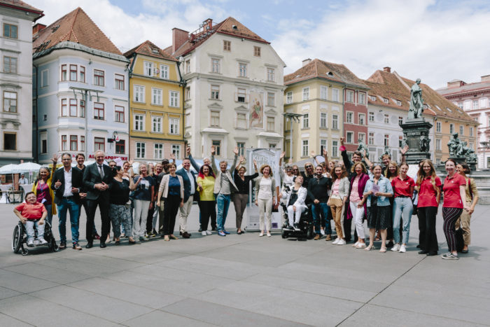 Auf einem Gruppenfoto stehen etwa 30 Personen in einem Halbkreis auf dem Grazer Hauptplatz. Im Hintergrund sind Häuser zu sehen. 