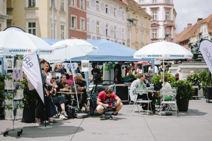 Auf dem Grazer Hauptplatz stehen viele Sonnenschirme, Menschen sitzen auf Stühlen oder stehen.