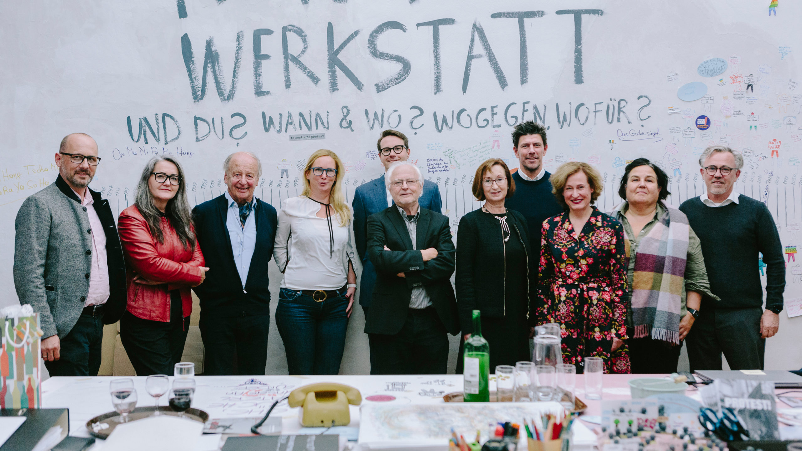 Die elf Mitglieder des Vorstandes des Vereins der Freund*innen des Graz Museums stehen nebeneinander vor einer Wand. Vor ihnen steht ein Tisch mit einem alten Telefon, Stiften, einer Flasche, einem Glaskrug und Gläsern.
