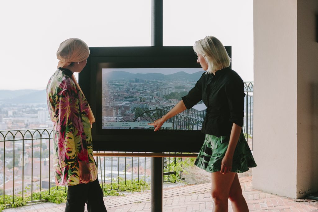 Zwei Frauen stehen vor der Sehmaschine in der Kanonenhalle des Graz Museum Schlossberg. Eine Frau zippt auf den Bildschirm.