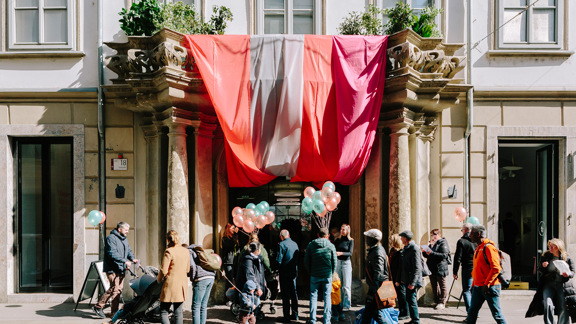 Vor dem Eingangsportal des Graz Museums in der Sackstraße tummeln sich viele Menschen. Vor dem Eingang werden Luftballons verteilt. Vom Balkon über dem Eingang hängen bunte Stoffe.