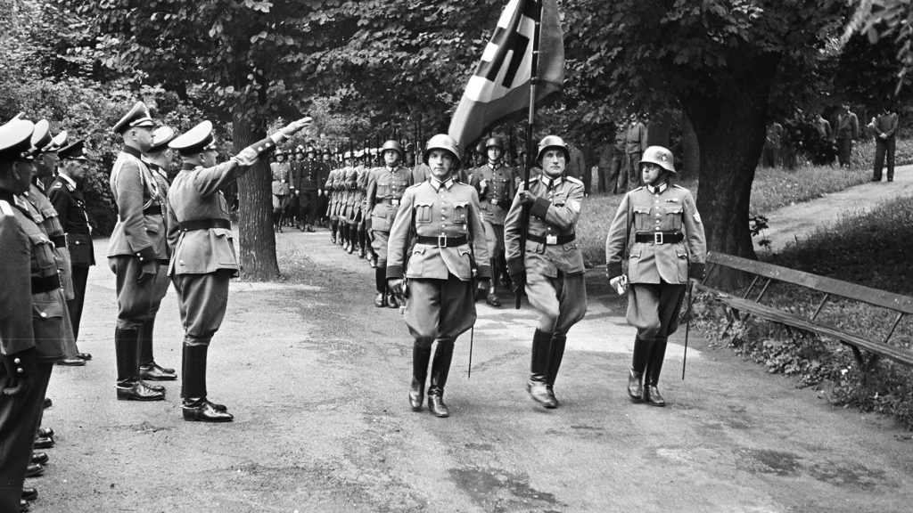 Auf dieser schwarz-weißen Fotografie marschieren Männer in Dreierreihen. Sie tragen Uniformen, Helme am Kopf und Lederstiefel. Der Mann in der ersten Reihe trägkt eine Fahne mit einem Hakenkreuz. Links von ihnen stehen weitere Männer in Uniformen. Einer von ihnen hebt den rechten Arm zum "Hitler-Gruß".