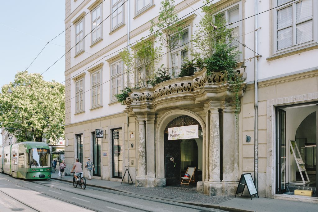 Das Eingangsportal des Graz Museums im Sommer. Auf dem Balkon über dem Eingang sind Pflanzen. Vor dem Gebäude fährt eine Straßenbahn vorbei.