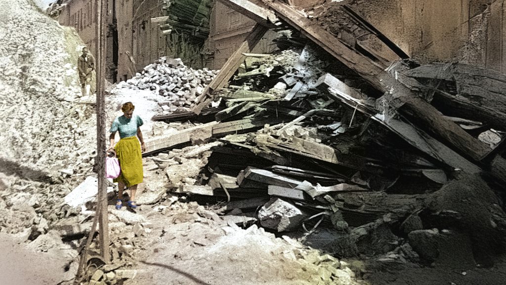 Eine Frau in einem bunten Kleid geht über ein Trümmerfeld aus zerstörten Gebäuden. Die trägt eine Tasche in der Hand. Hinter ihr geht ein Mann.
